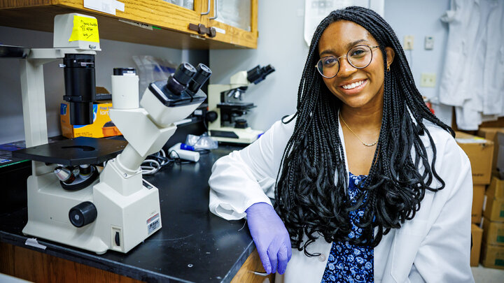 Laila Doyle, a senior nutritional science and dietetics major at the University of Nebraska–Lincoln, poses next to two microscopes in a lab. She is wearing a white labcoat over a blue floral dress, with purple protective gloves and glasses.